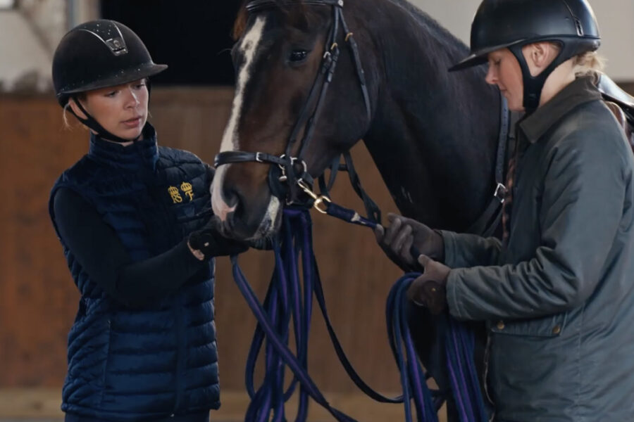 Två personer i ridhjälm leder och hanterar en sadlad häst i ett stall