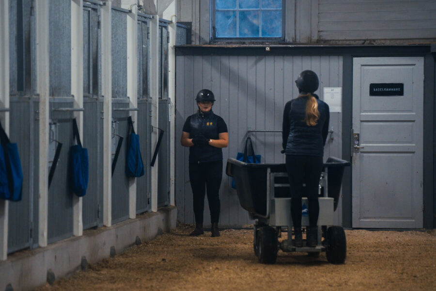 Två personer i ridhjälm i ett stall, en står bredvid en vagn på stallgången