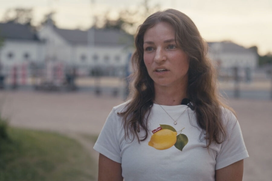 Kvinna utomhus i närbild med vit tröja med citrontryck och mikrofon, framför suddiga byggnader i skymning
