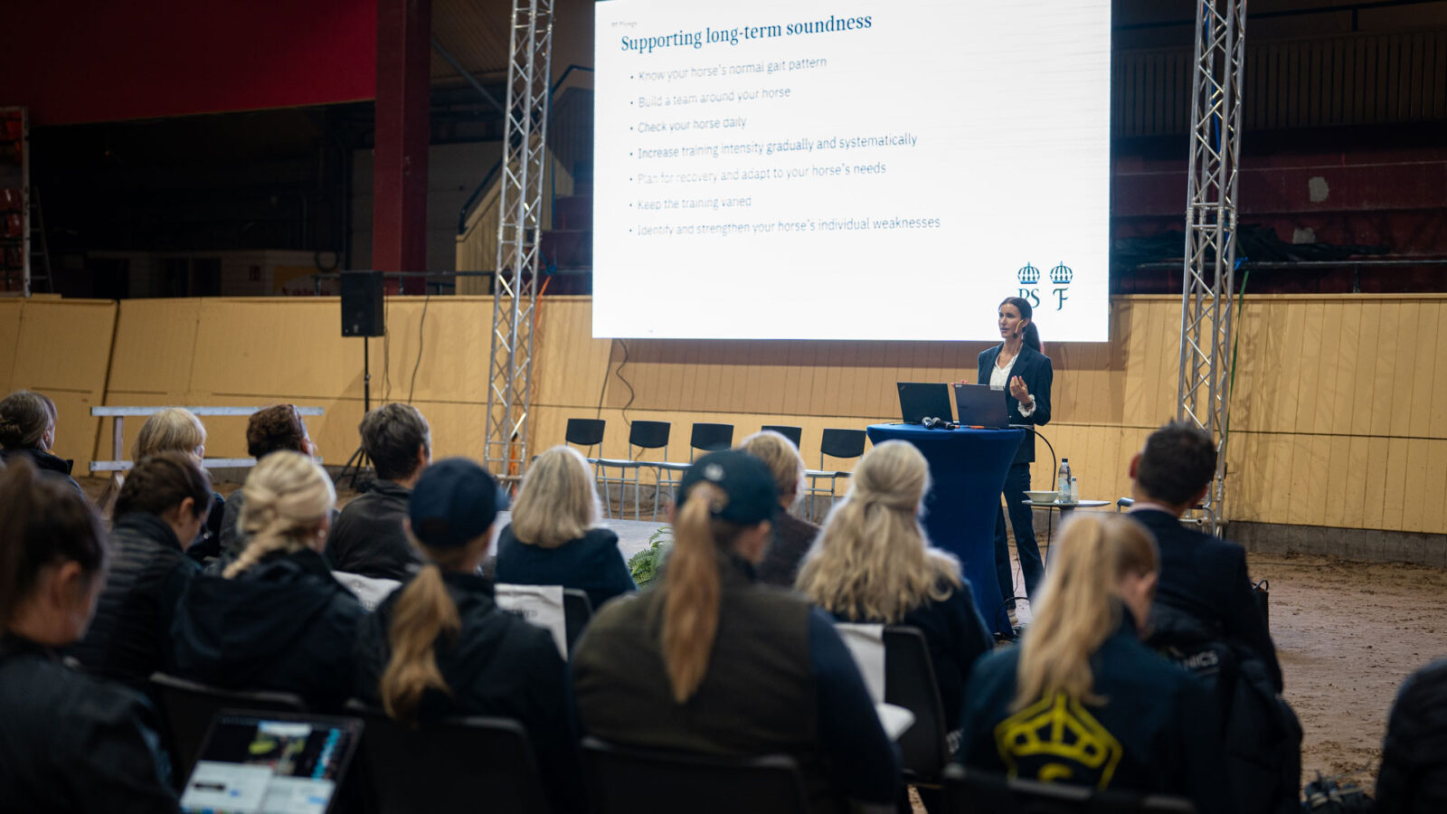 Kvinna håller föredrag inför publik i ett ridhus, med en projicerad presentation om hästars långsiktiga hälsa