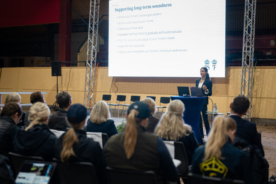 Kvinna håller föredrag inför publik i ett ridhus, med en projicerad presentation om hästars långsiktiga hälsa