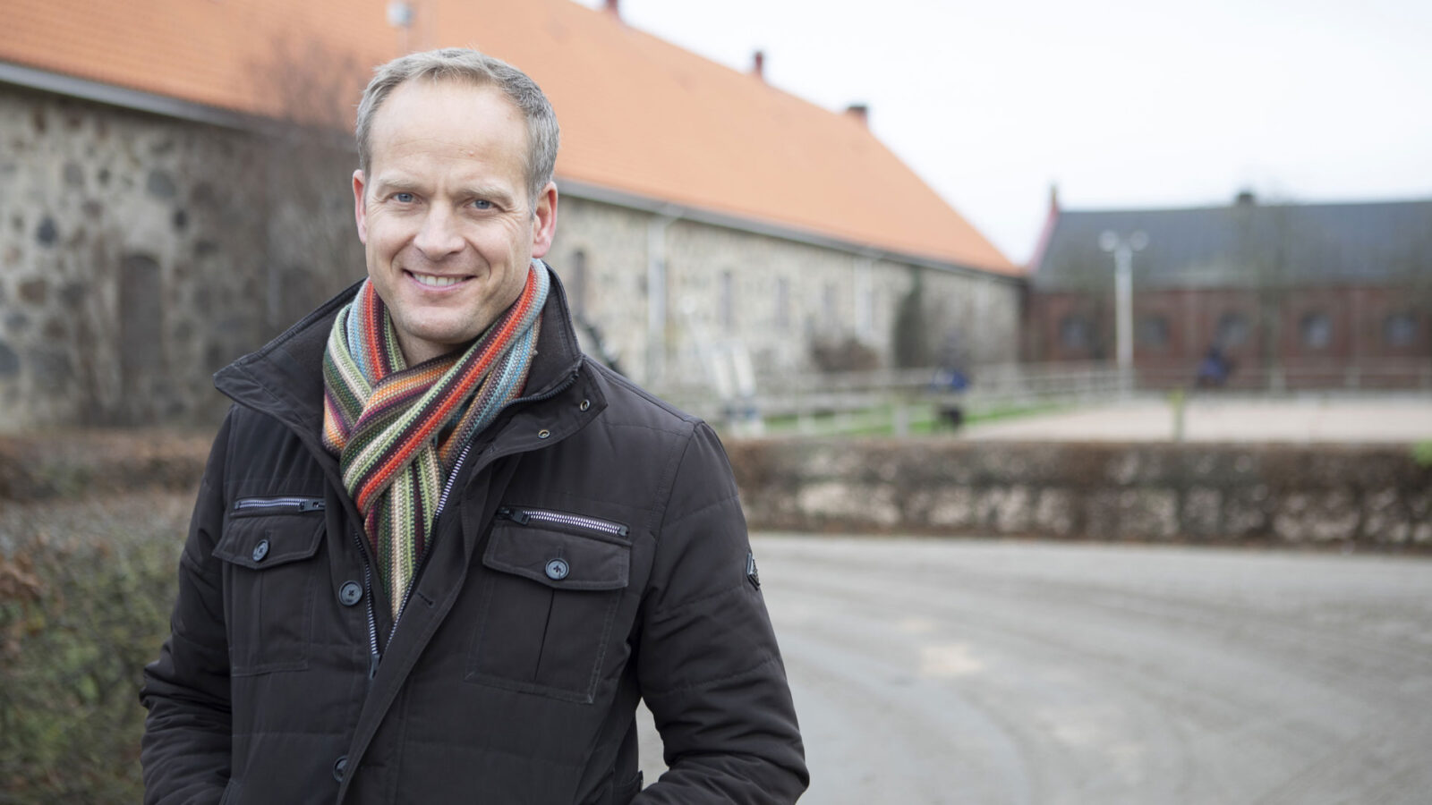 Leende man i vinterjacka och randig halsduk framför byggnader på landet