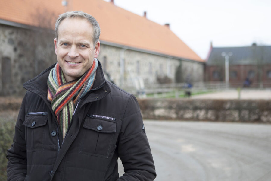 Leende man i vinterjacka och randig halsduk framför byggnader på landet