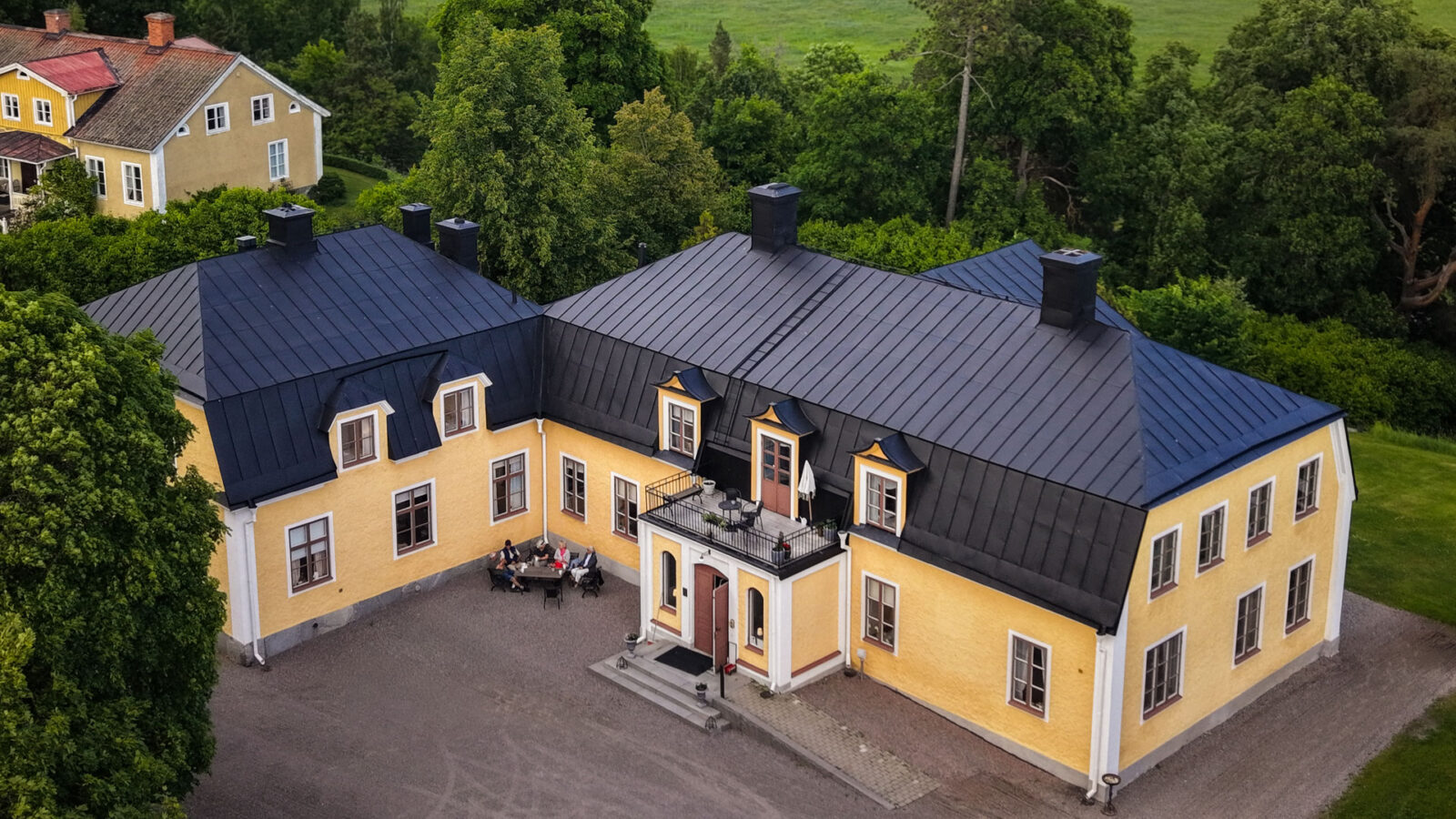 Flygbild över en gul herrgård med svart tak, omgiven av träd och grönska