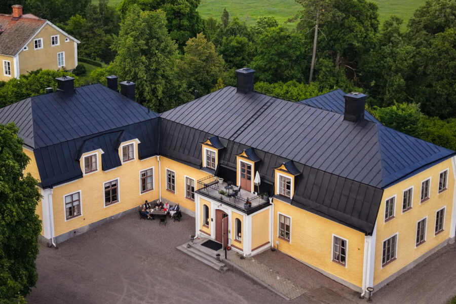 Flygbild över en gul herrgård med svart tak, omgiven av träd och grönska
